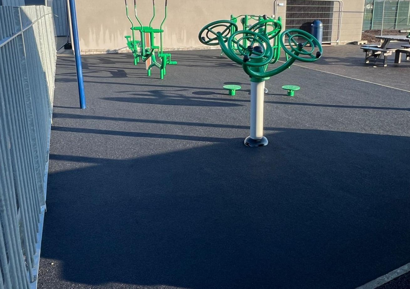 Green outdoor gym equipment on a charcoal rubber crumb surface, with the school building in the background